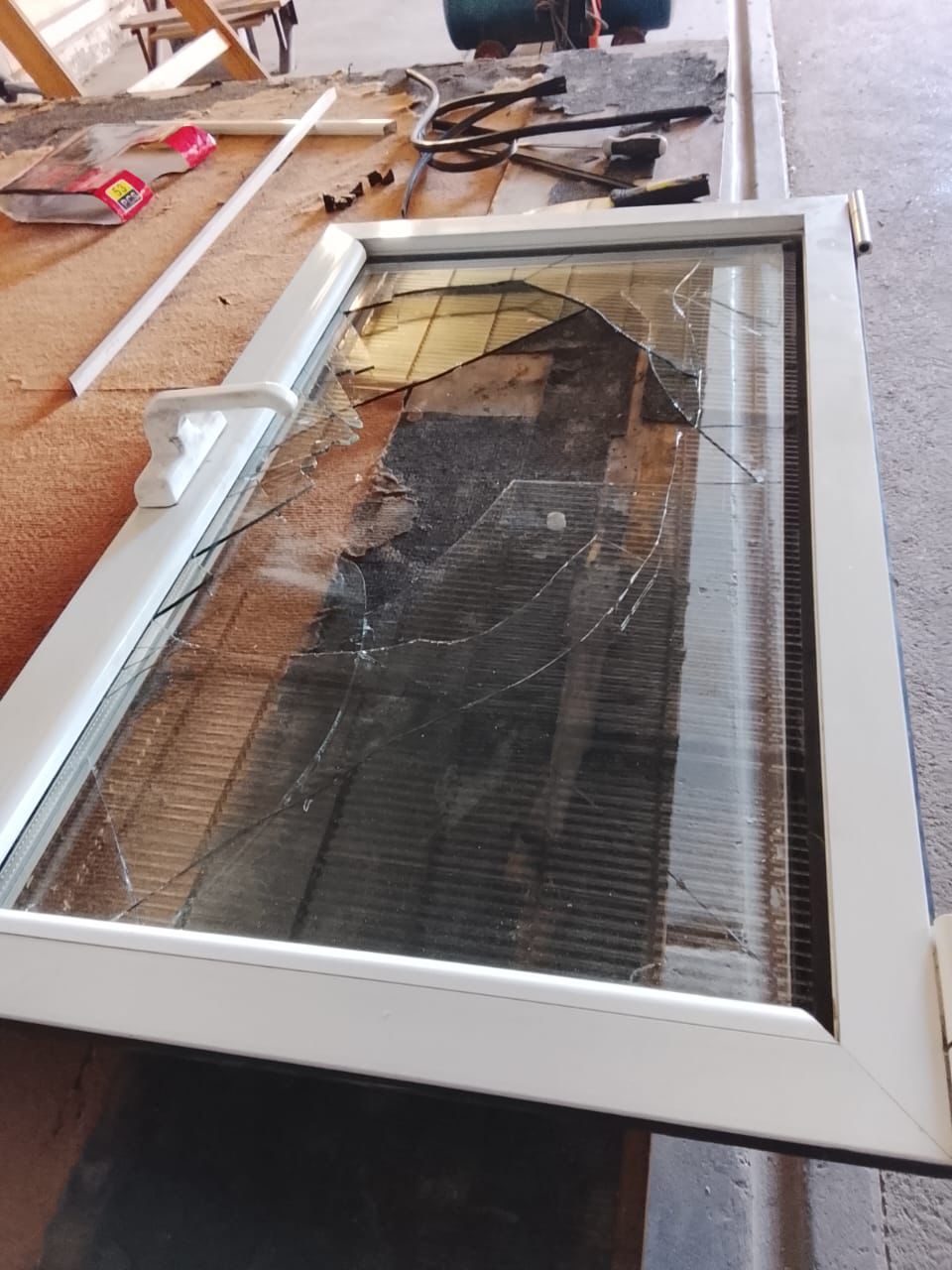 Cristal de ventana roto, con marco blanco, sobre una superficie de madera y con herramientas esparcidas por todas partes.