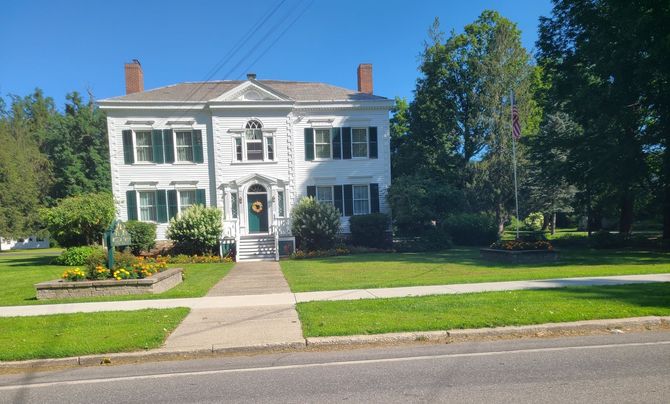 White two-story colonial house with columns, front lawn, garden, and trees on a sunny day