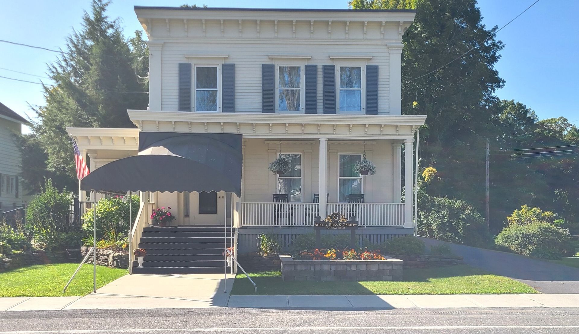 Two-story white house with blue shutters, front porch, and black awning, viewed from the street