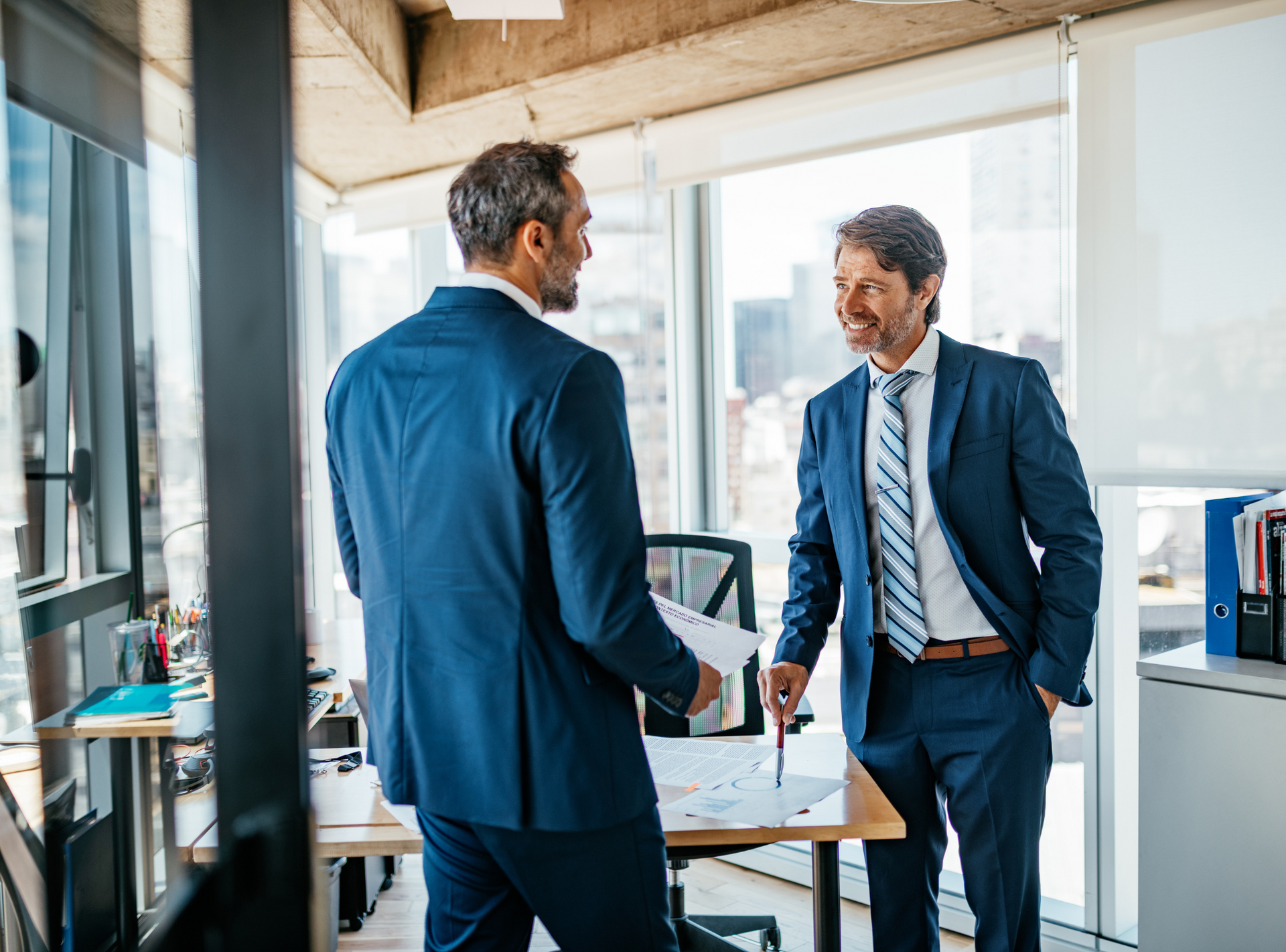 Two men in suits and ties are standing in an office talking to each other.