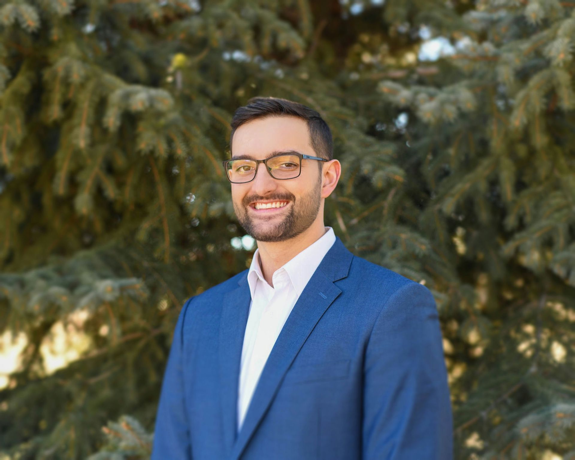A man in a suit and glasses is standing in front of a tree.
