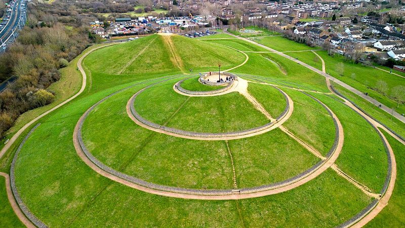 Green, spiral-shaped hill with pathways. A monument stands on top. Houses and a road are in the background.