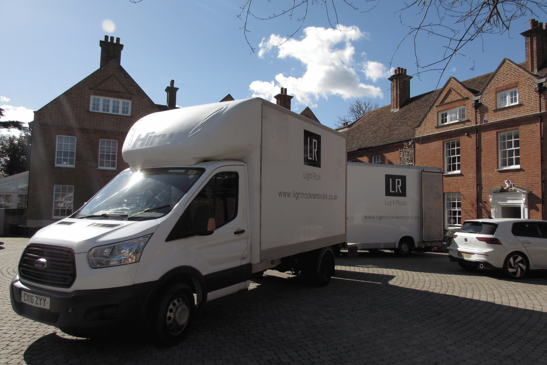 Two white moving trucks parked in front of a brick building; sunny day.
