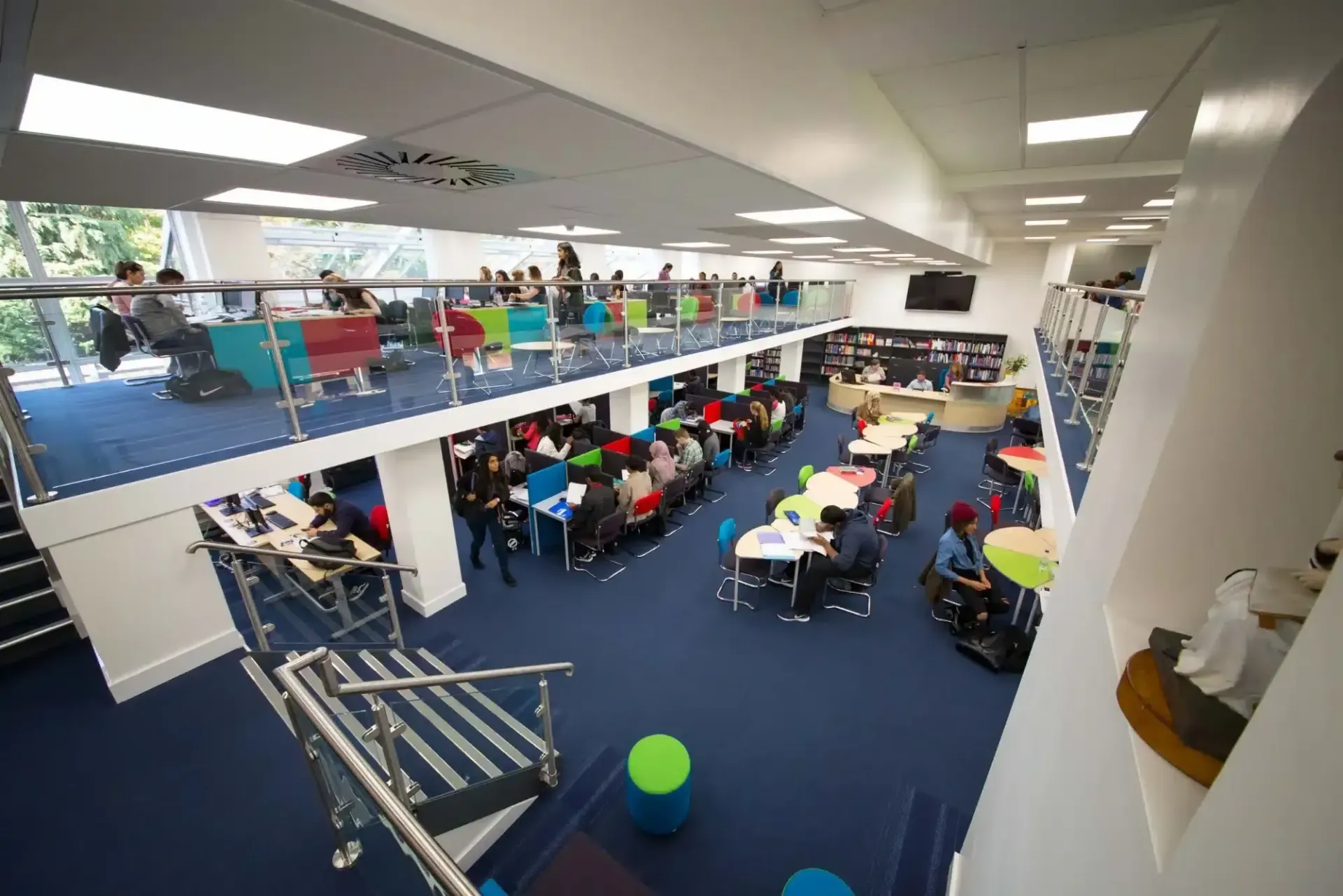 A two-story modern library with students studying at tables, blue carpet, and colorful seating.