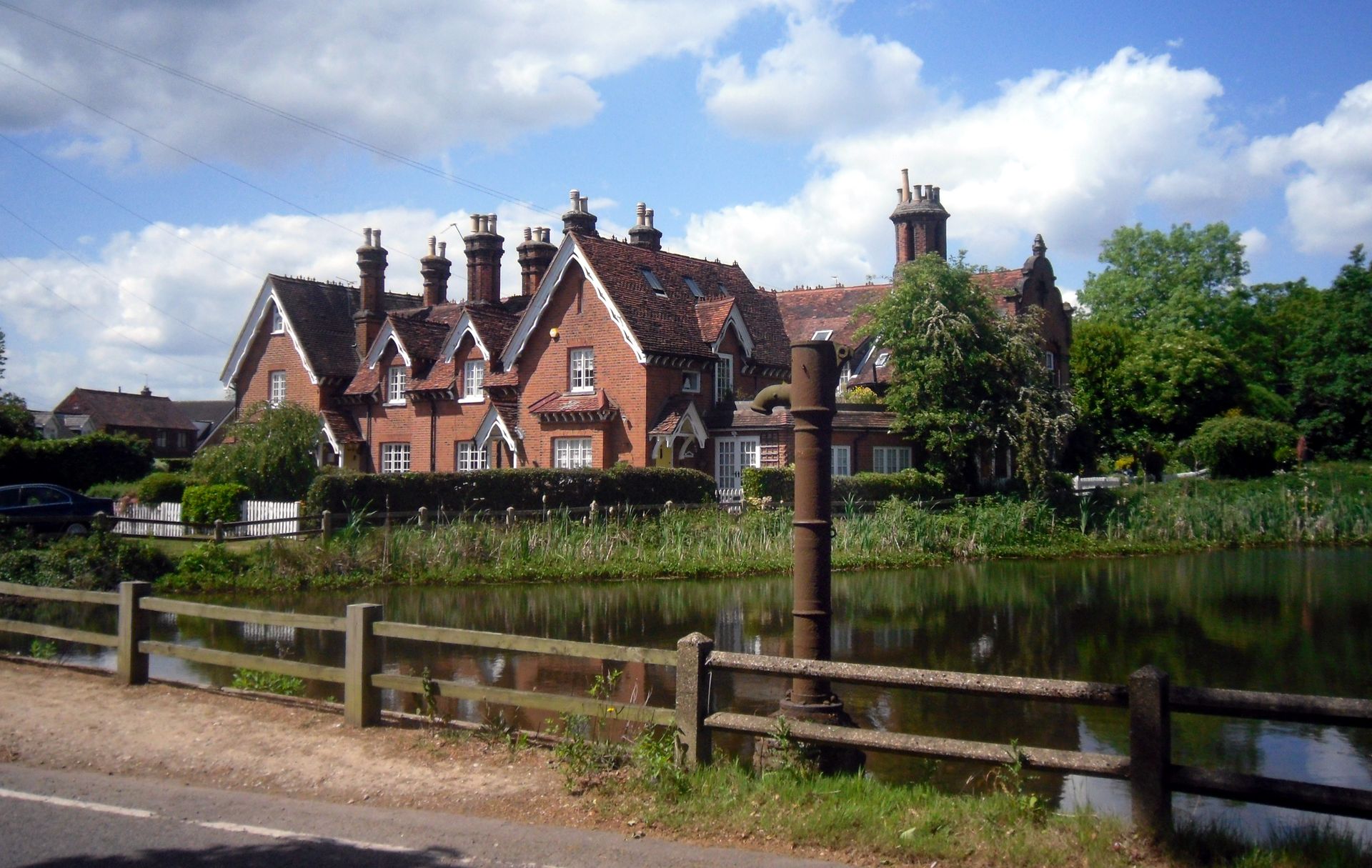 Red brick houses with chimneys, beside a pond and wooden fence, under a blue sky.