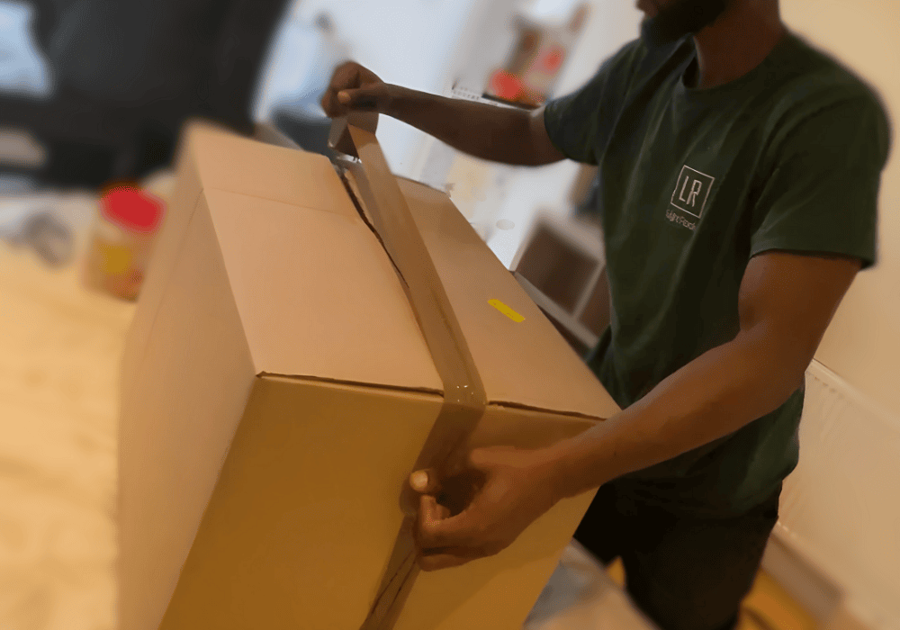 A person taping a cardboard box shut. They wear a blue shirt and tan pants. Other boxes are in the background.