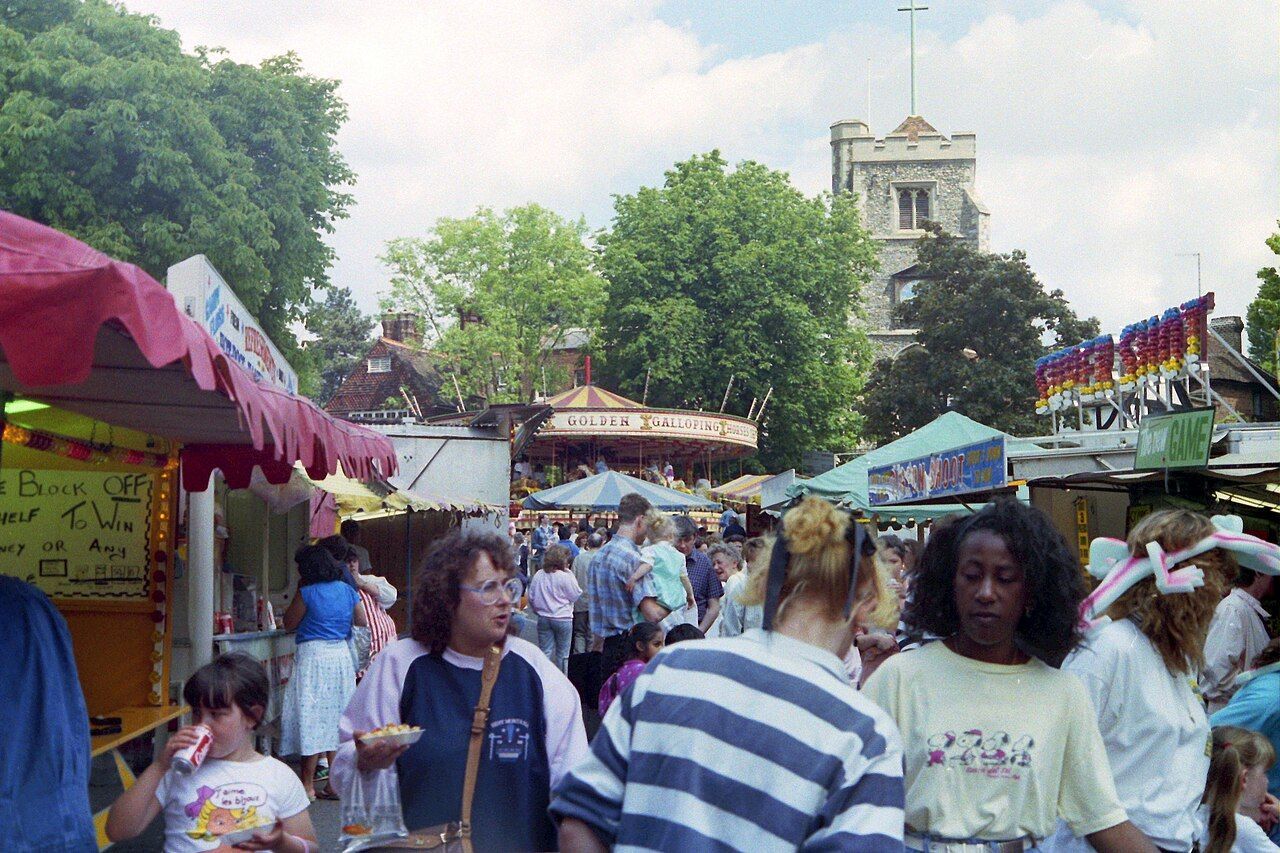 Fairground scene with crowds, food stalls, and a carousel, church tower in the background.