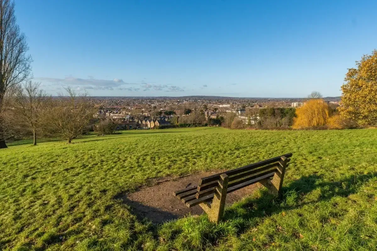 Wooden park bench on a grassy hill overlooking a distant cityscape under a clear blue sky.