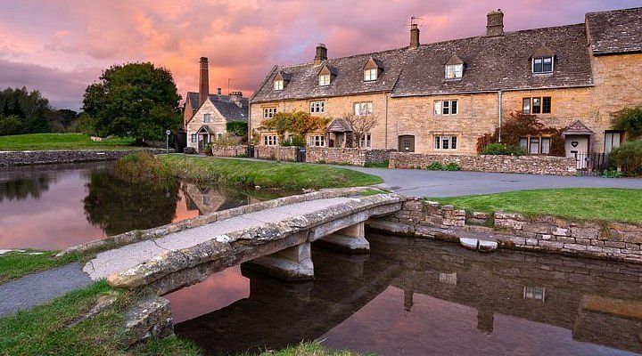 Stone cottages line a river in a Cotswold village, a small bridge in the foreground reflecting the colorful sunset sky.