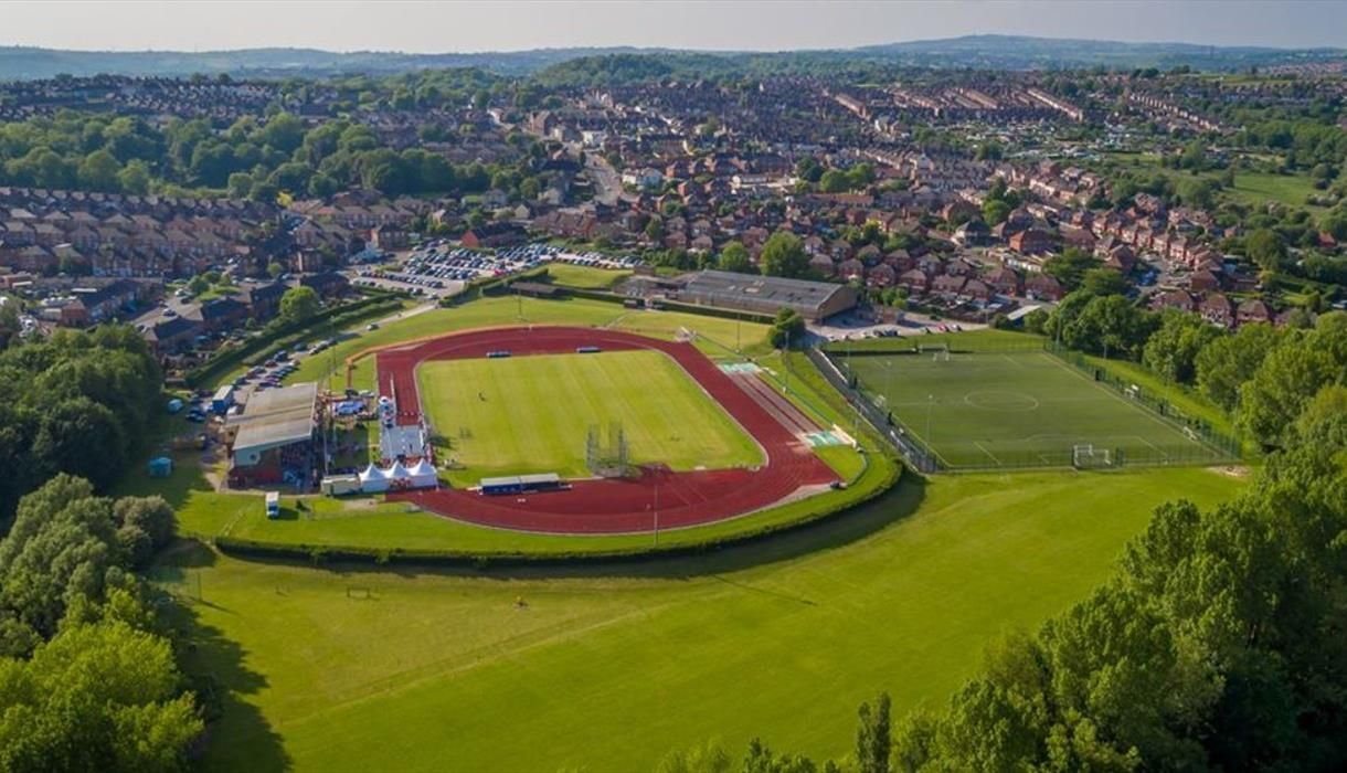 Aerial view of a sports complex with a red running track and green soccer field next to green grassy areas and trees.