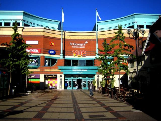 St. George's Shopping Centre entrance with blue accents, flags, and a pedestrian walkway on a sunny day.