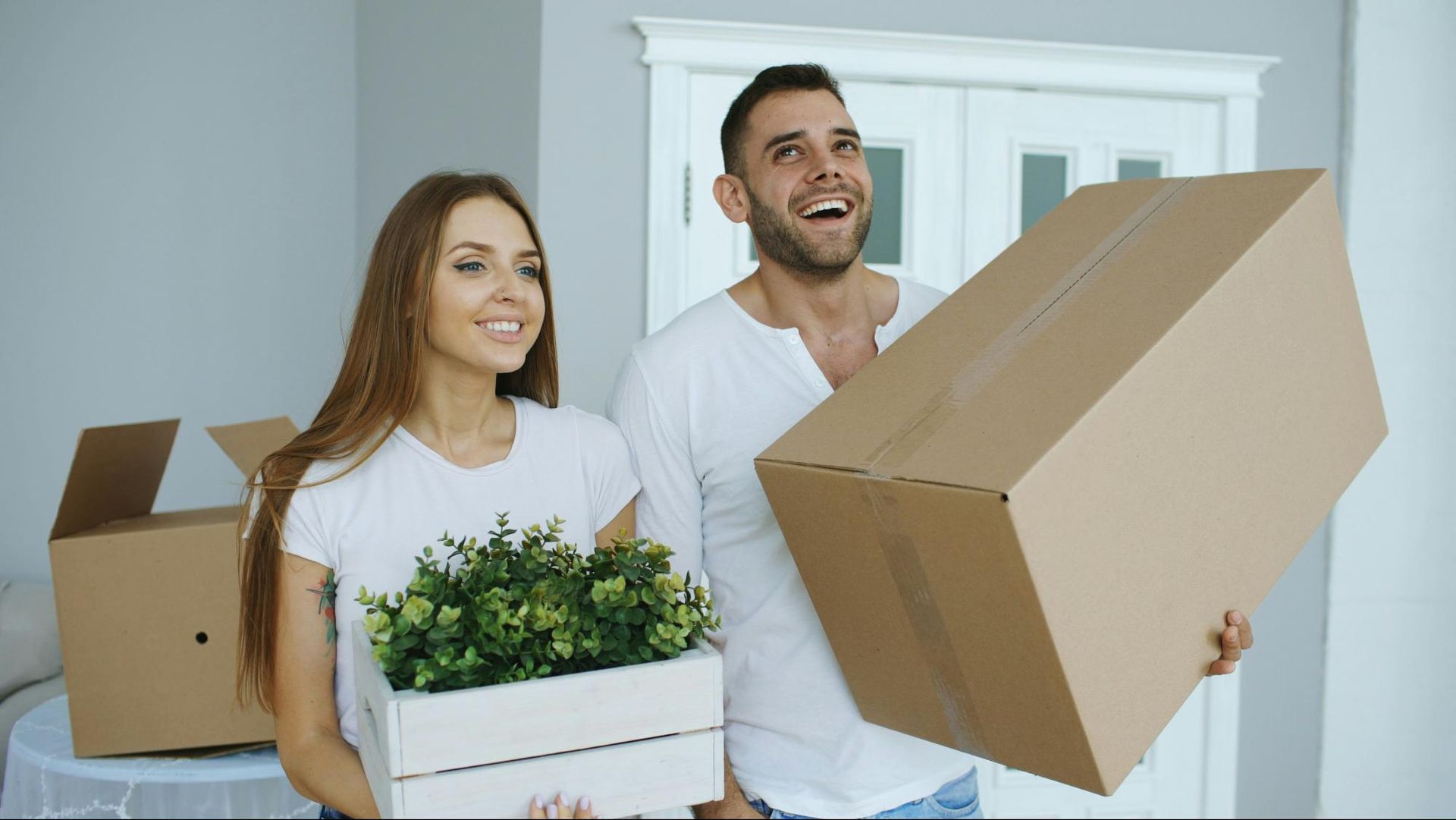 Couple moving into a new home, carrying boxes and plants, smiling.