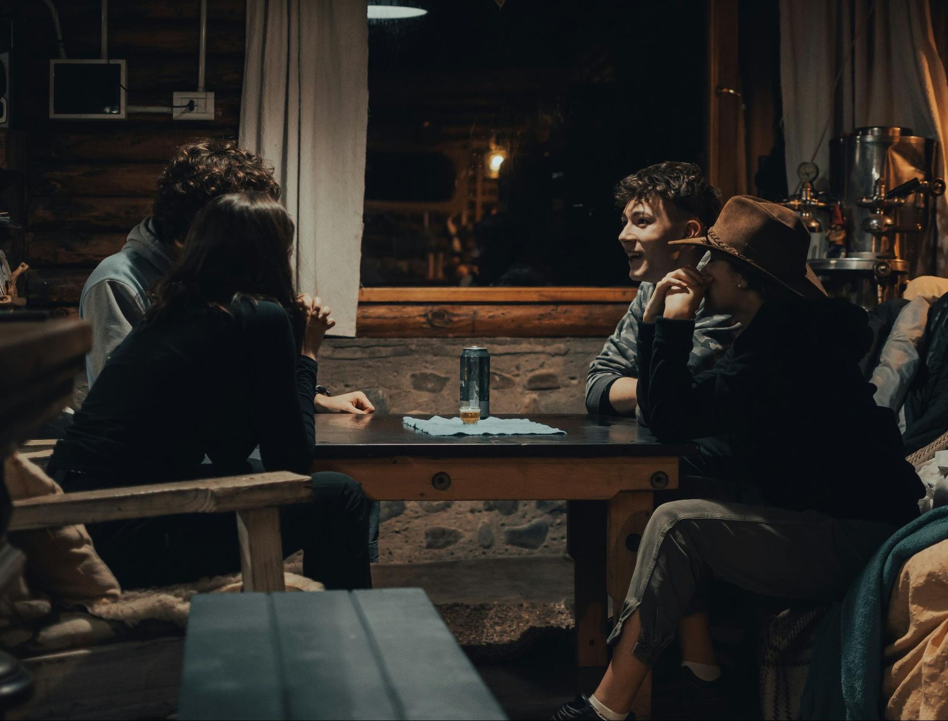 Four people seated around a table playing cards inside a dimly lit cabin. One smiles broadly.