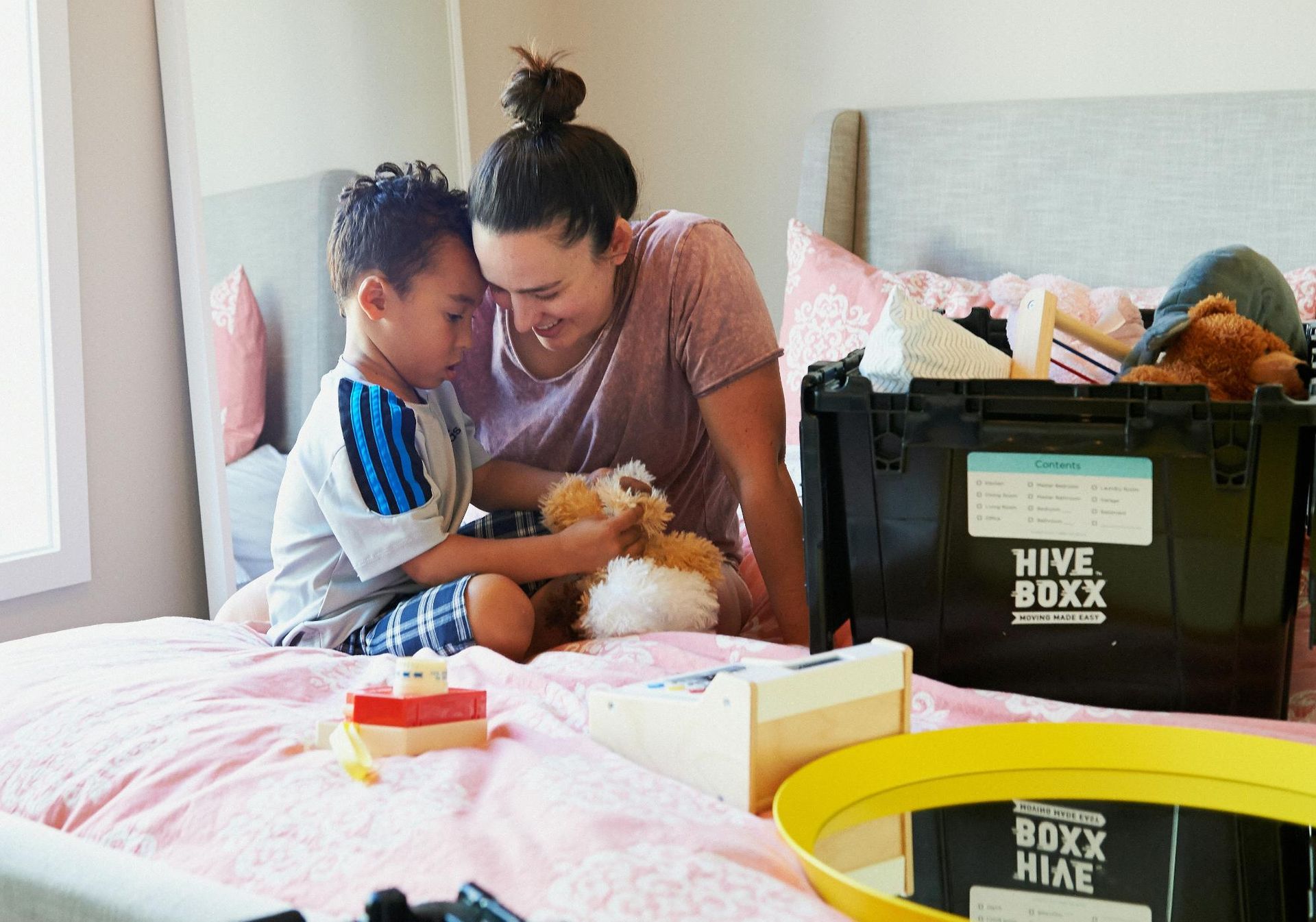 Woman and child on a bed, packing belongings. Child holds a stuffed animal, both smiling. A black storage bin is nearby.