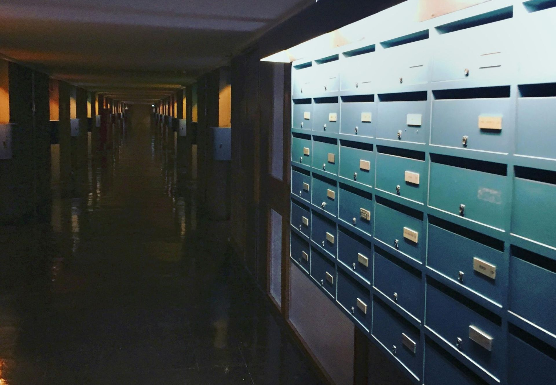 Long, dimly lit hallway with mailboxes on the right. Blues, greens, and browns dominate the color scheme.