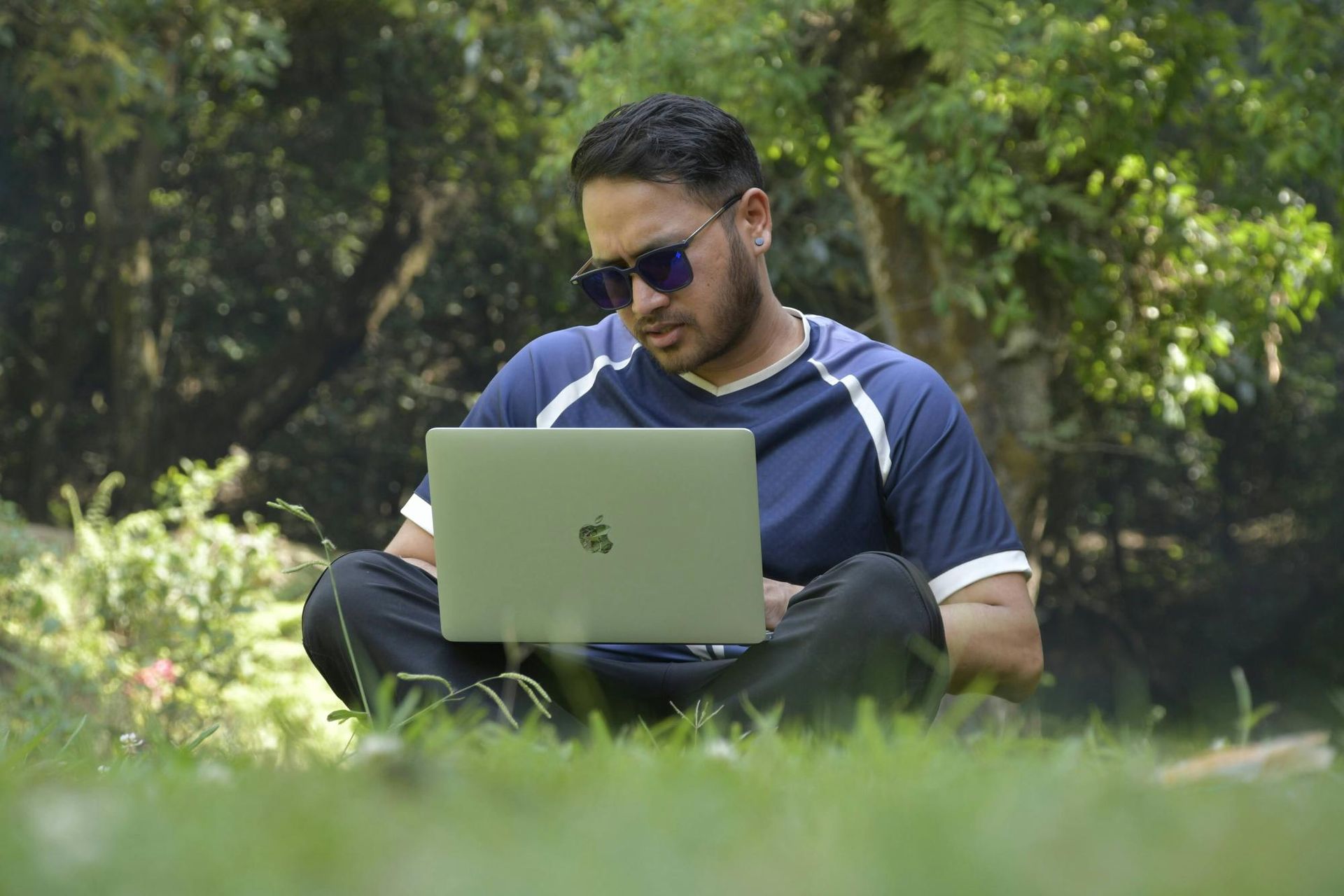 Man in sunglasses sits cross-legged on grass, using a laptop in a park setting with trees.