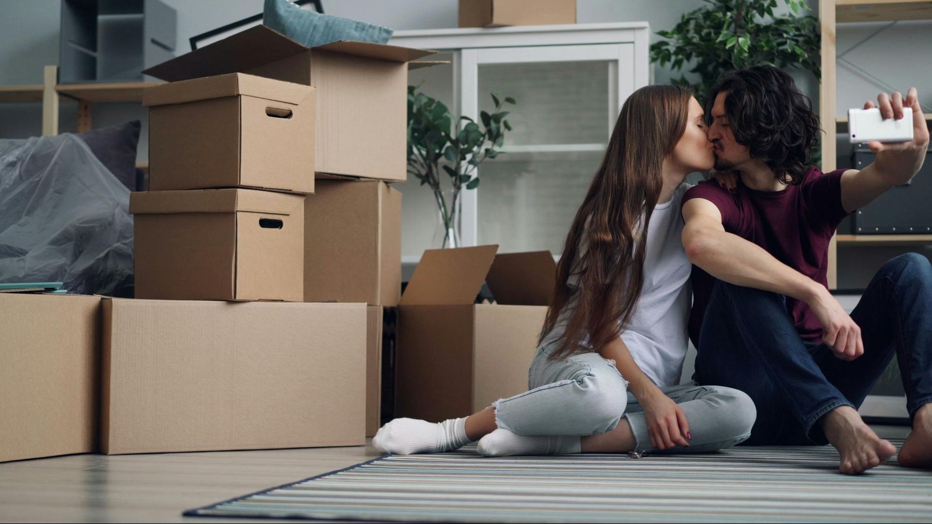 Couple kissing and taking a selfie while sitting on floor; surrounded by moving boxes.