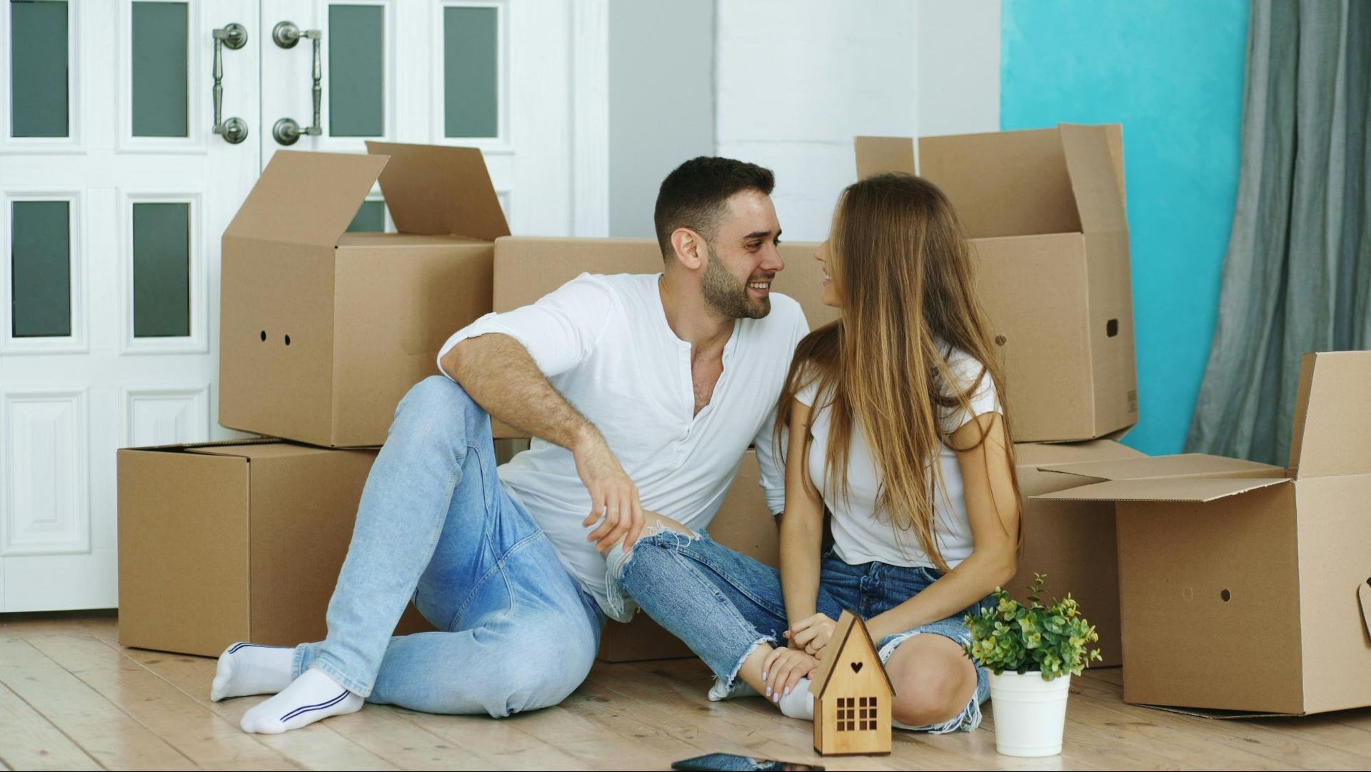 Couple sitting on the floor with moving boxes, smiling and looking at each other in a new home.