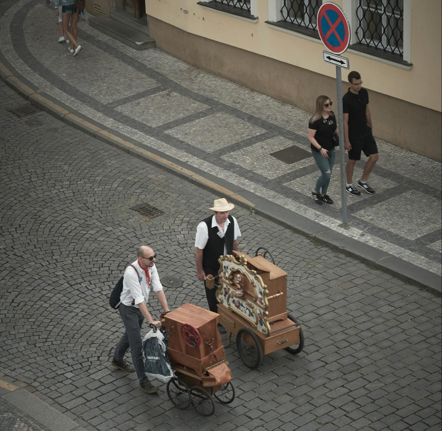 Two men push antique music boxes on a cobblestone street, near pedestrians.