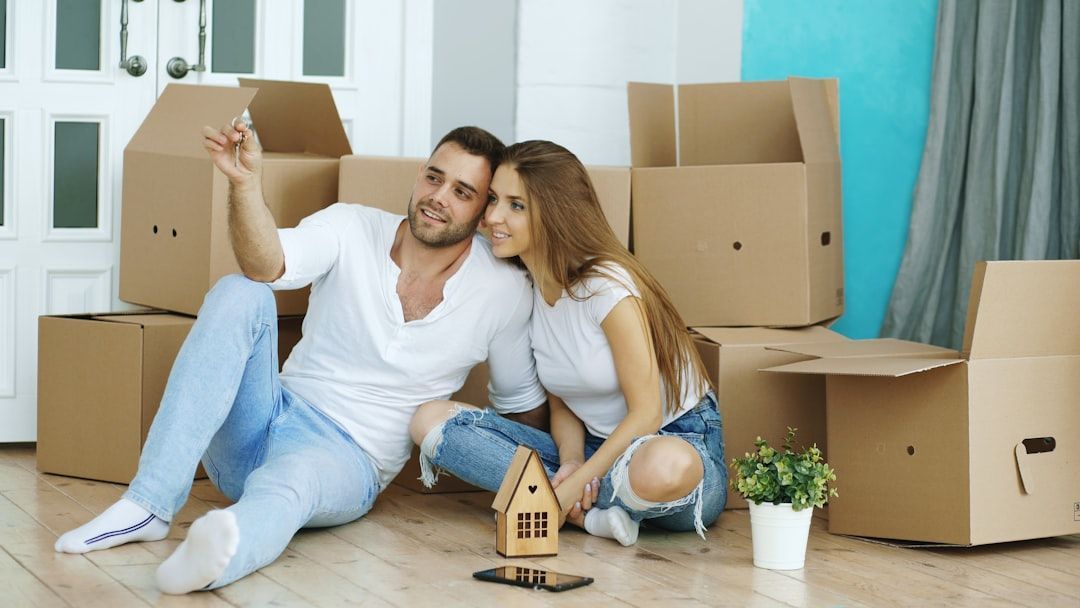 Couple sitting on floor, surrounded by moving boxes, man pointing, smiling woman.