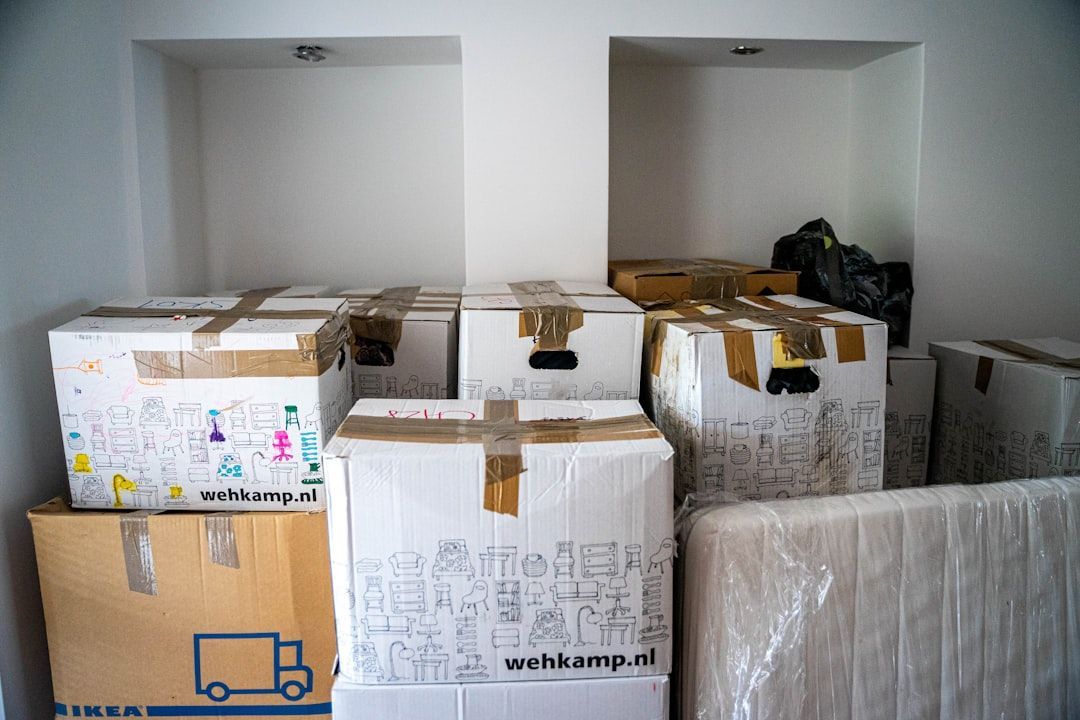 Cardboard moving boxes stacked in a white-walled storage niche. Some boxes sealed with tape, one with doodles.