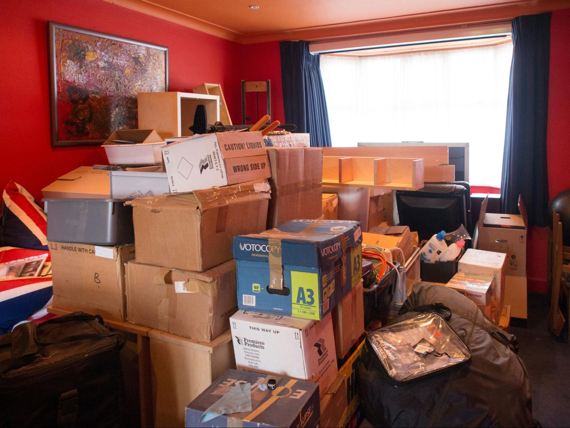 Room filled with cardboard boxes, possibly moving. Red walls, window with dark curtains, and a painting visible.