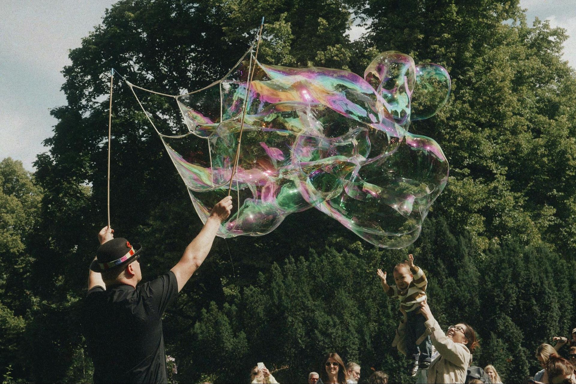 Man makes giant bubble wand. Bubbles float in air. Child reaches toward them. Outdoors, trees in background.