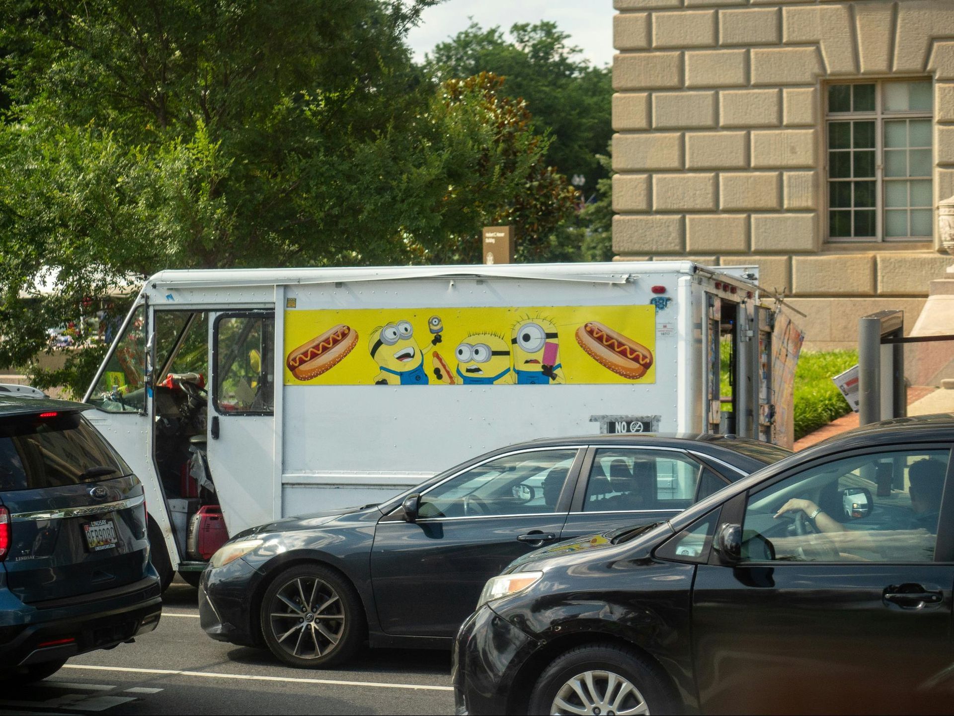 Hot dog food truck with Minion characters parked near a brick building. Cars are in front.