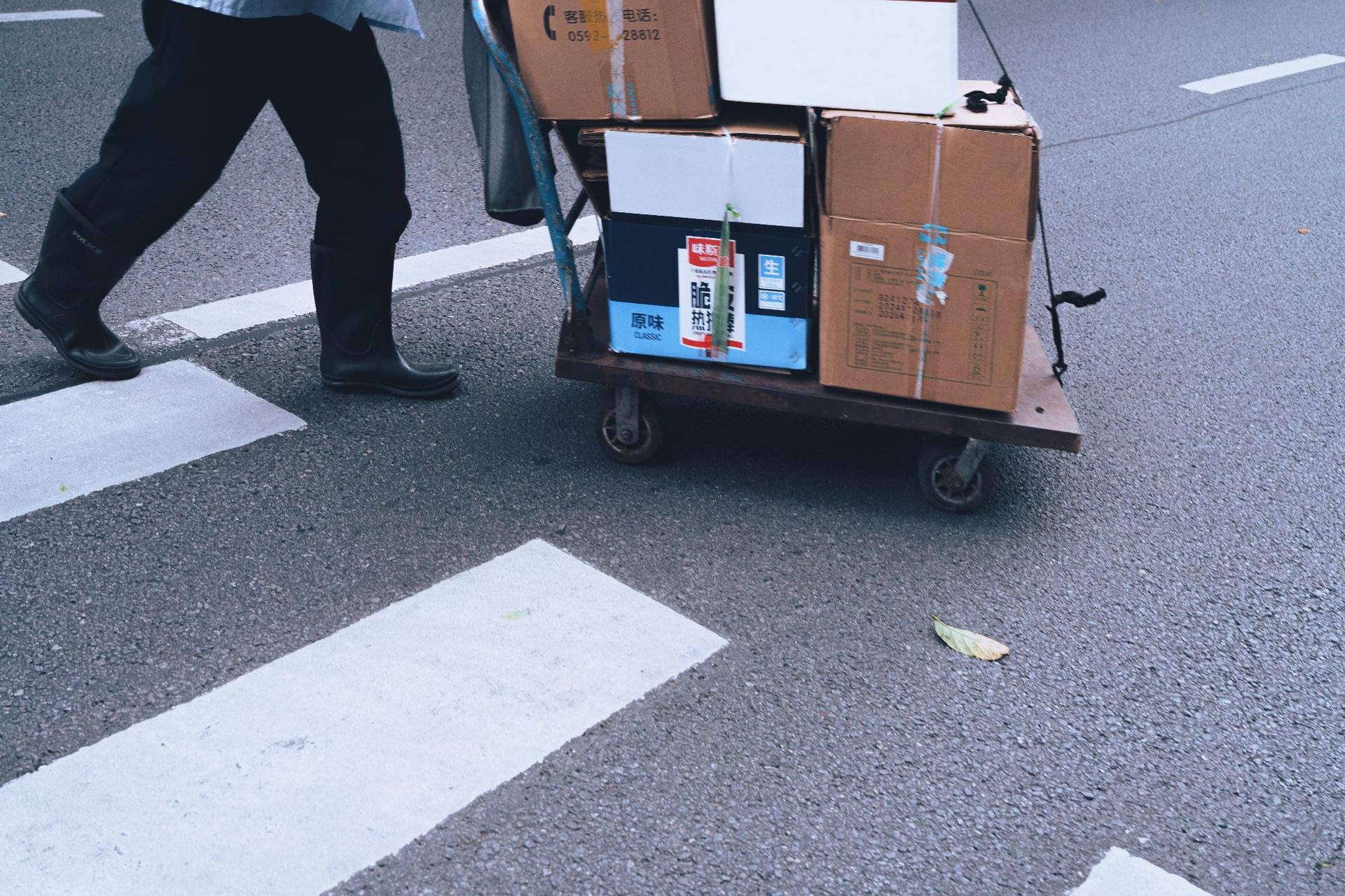 Person pulling a cart loaded with boxes across a crosswalk. Asphalt road with white painted stripes.