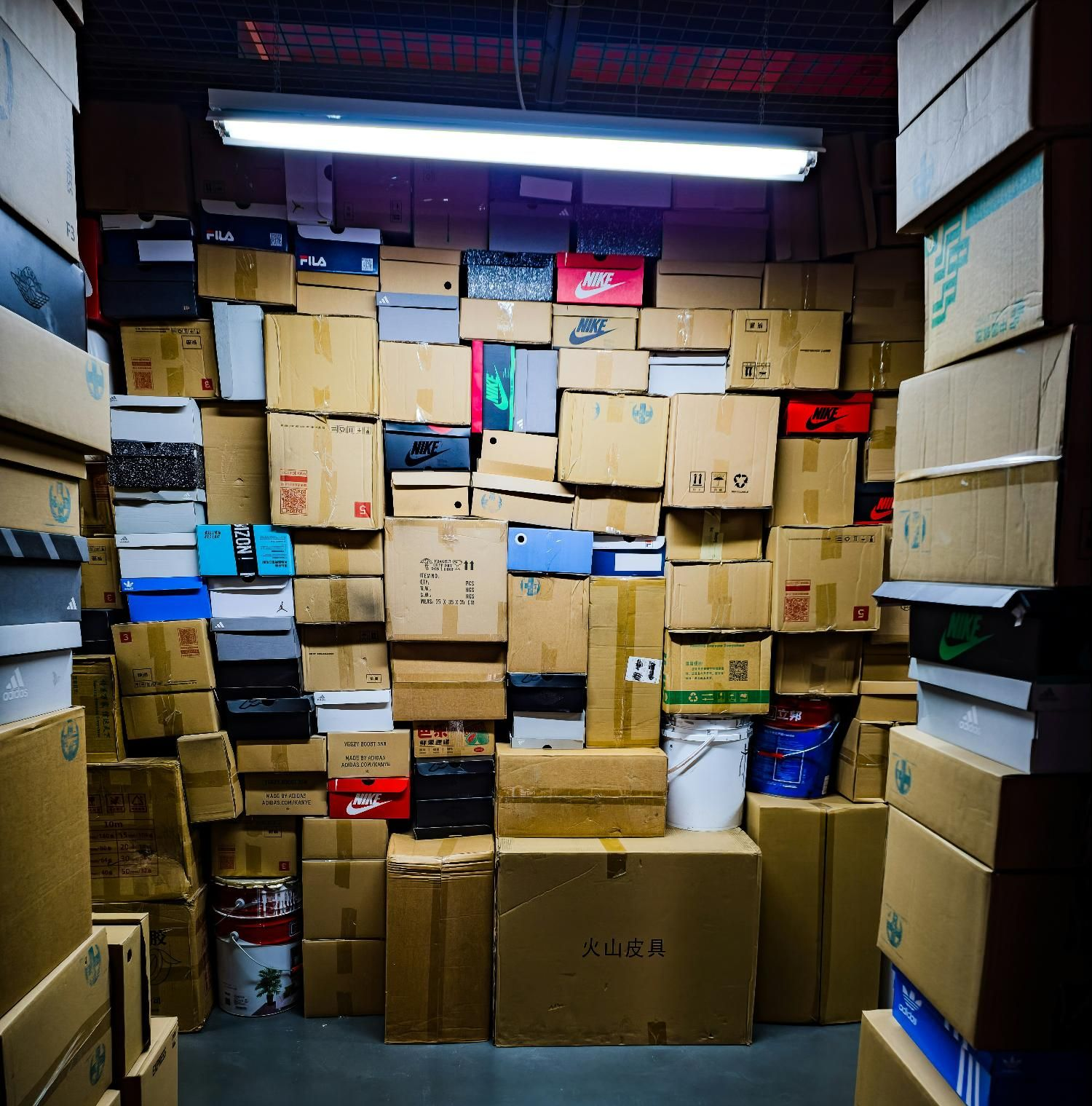 A storage unit overflowing with stacked cardboard boxes, many shoe boxes, lit by a fluorescent ceiling light.