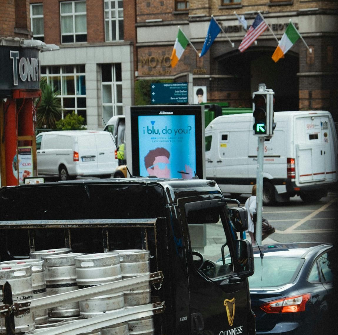 Street scene with trucks, vans, and a green traffic light. Buildings with flags. An illuminated advertisement.