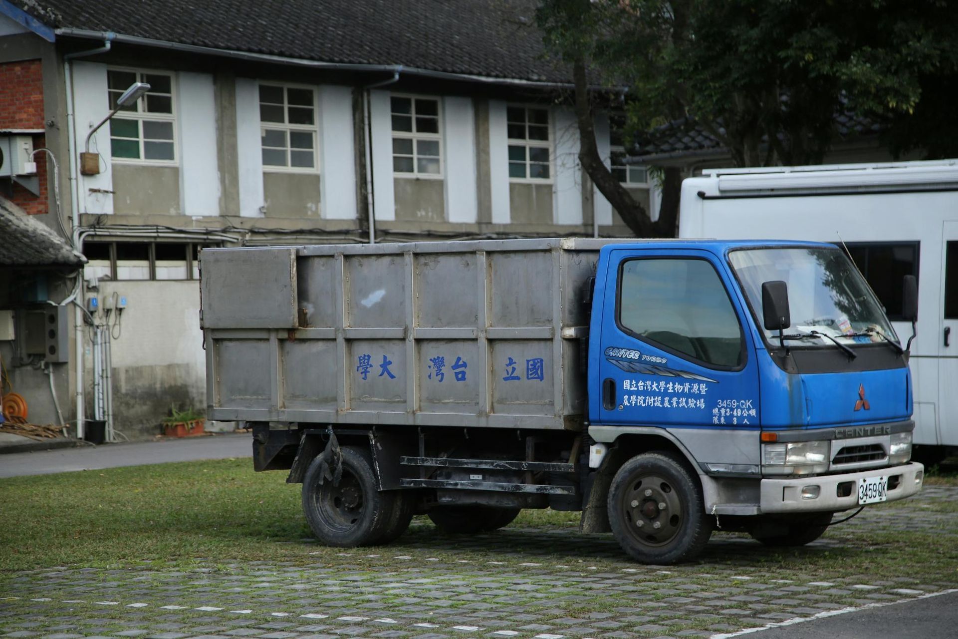 Blue and silver dump truck parked on grass, in front of a building with windows.