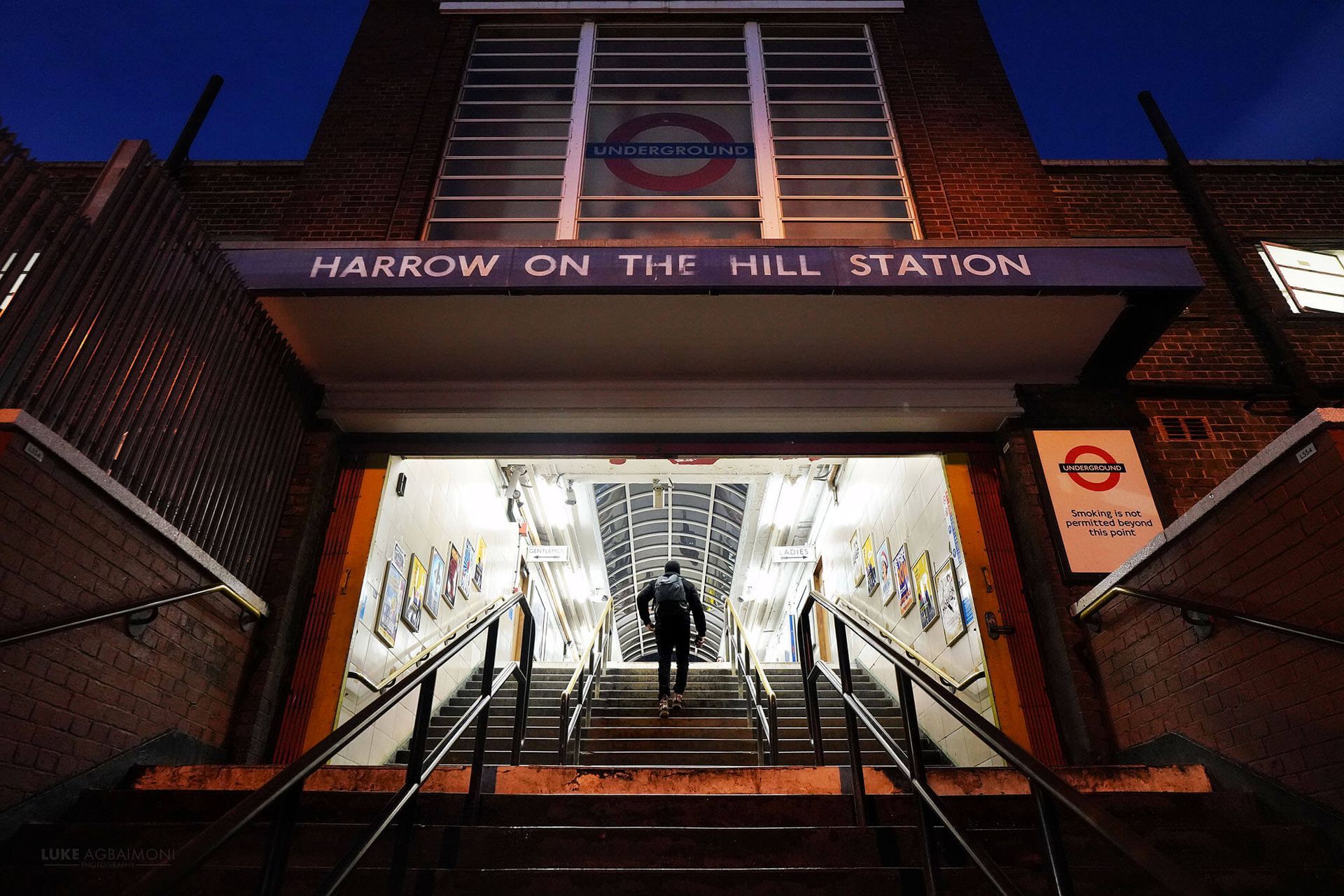 Entrance to Harrow on the Hill tube station at dusk; a person walks down the stairs into the station.
