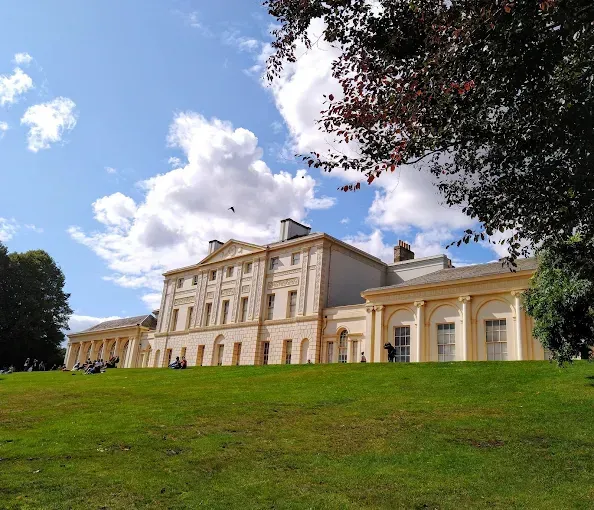 Large light-colored building atop a grassy hill under a blue sky with puffy clouds.