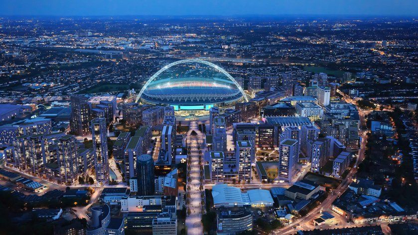 Aerial view of Wembley Stadium lit up at night, surrounded by modern buildings and city lights under a twilight sky.