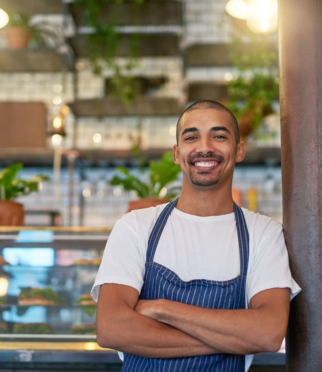 A man in an apron is standing in a restaurant with his arms crossed and smiling.