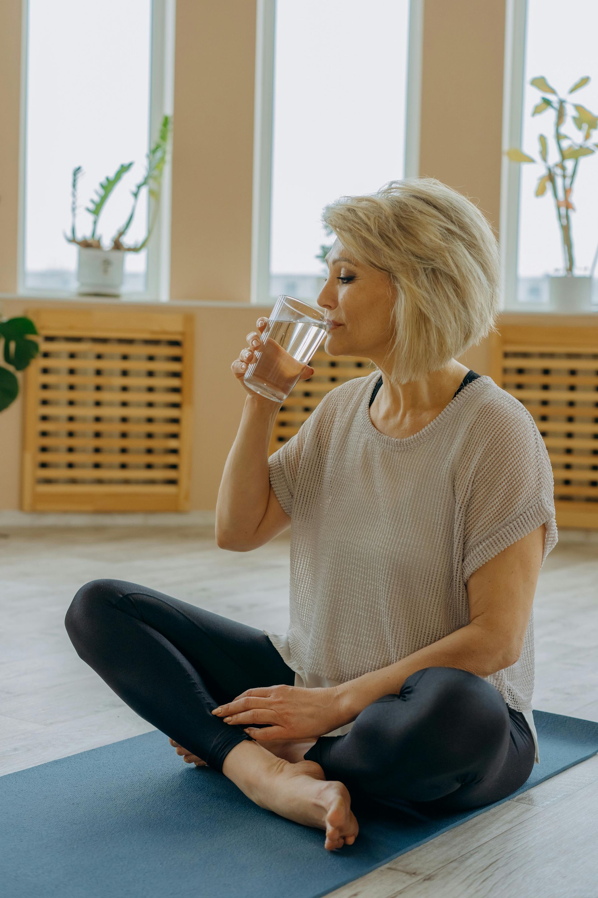 A woman is sitting on a yoga mat drinking a glass of water.