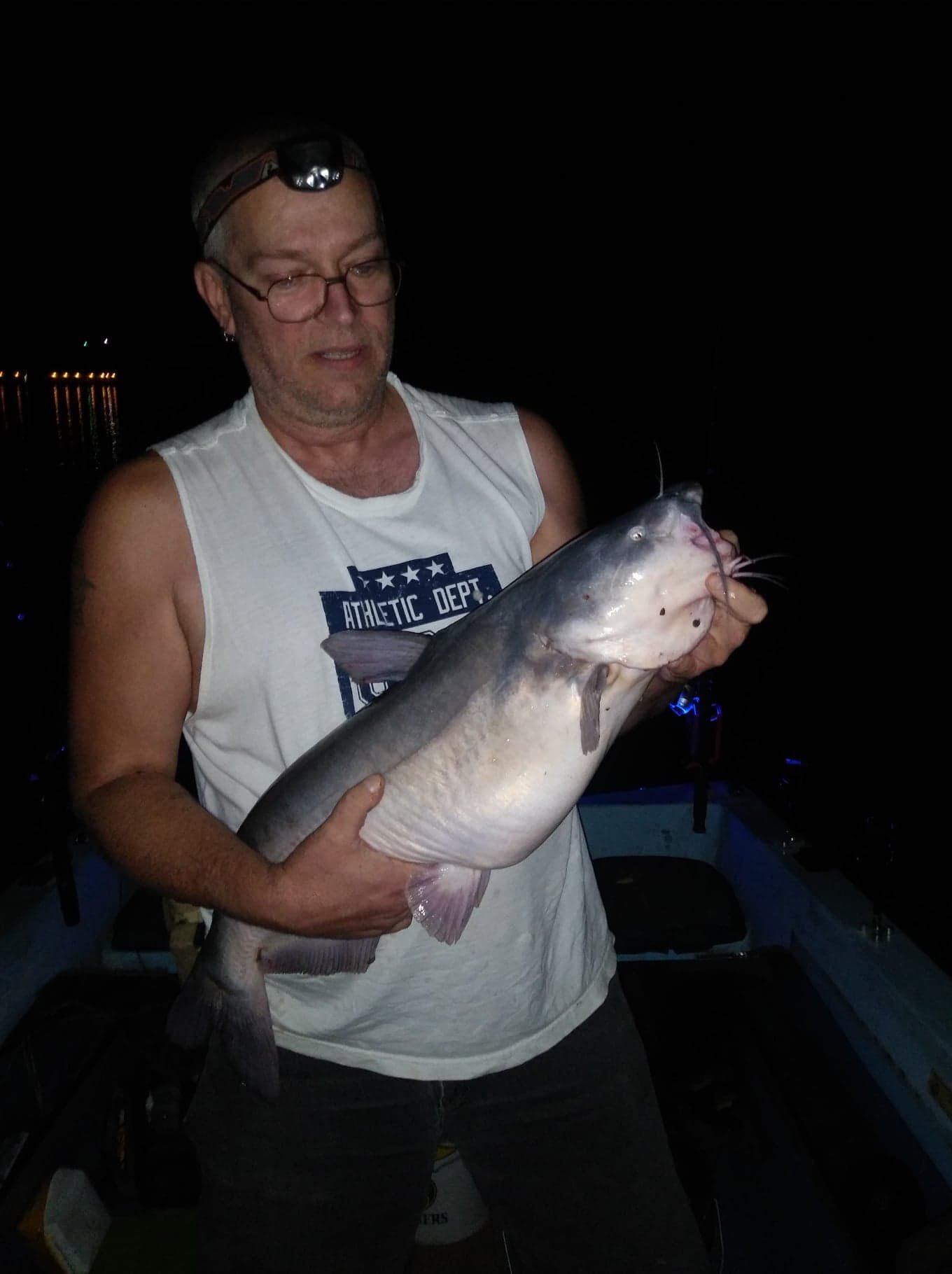 Man holding a large, gray catfish in a boat at night.