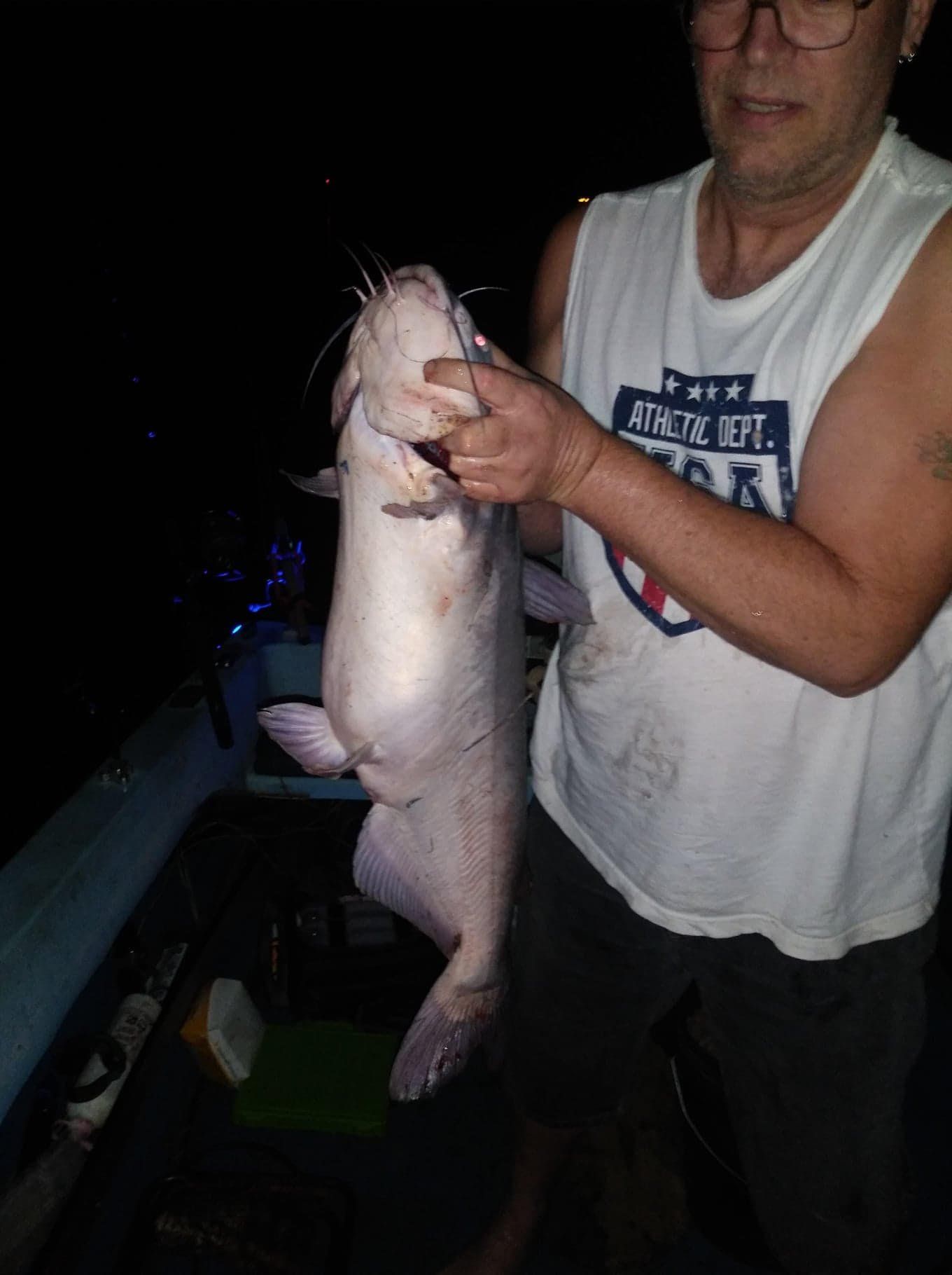 Man holding a large catfish at night, possibly on a boat.