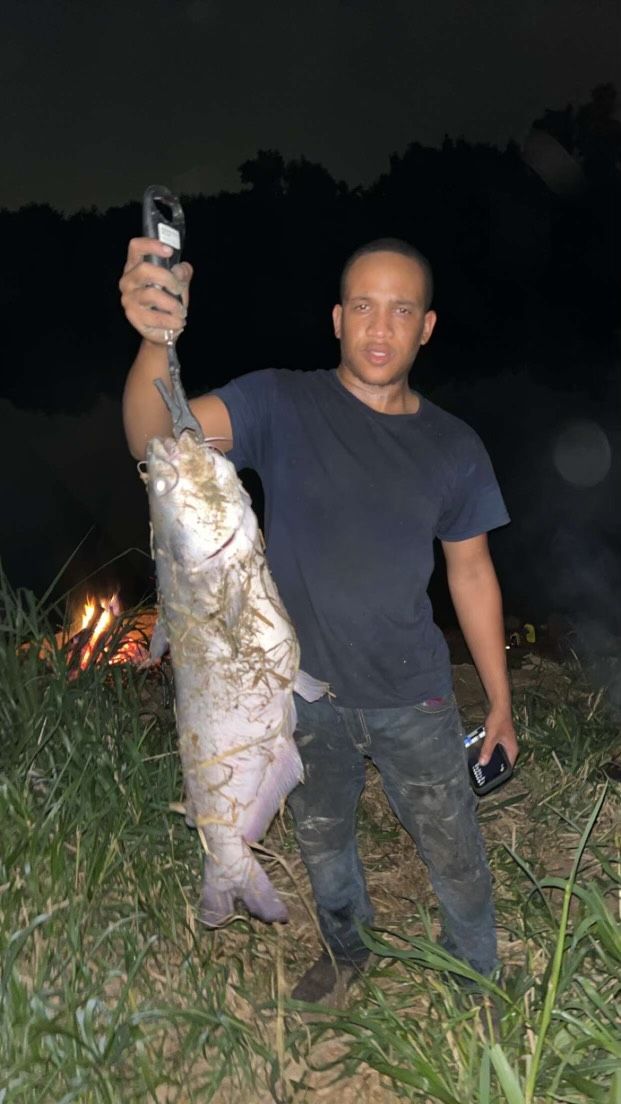 Person holding up a large catfish; night setting with a fire burning.