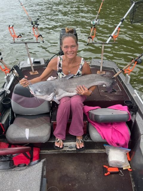 Woman in a boat holding a large catfish. Several fishing rods are visible.