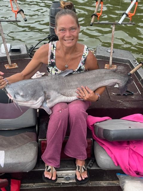 Woman seated in a boat holding a large blue catfish, smiling.