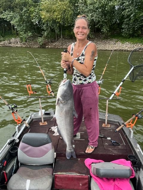 Woman on a boat holding a large fish, fishing rods visible. River setting, cloudy sky.
