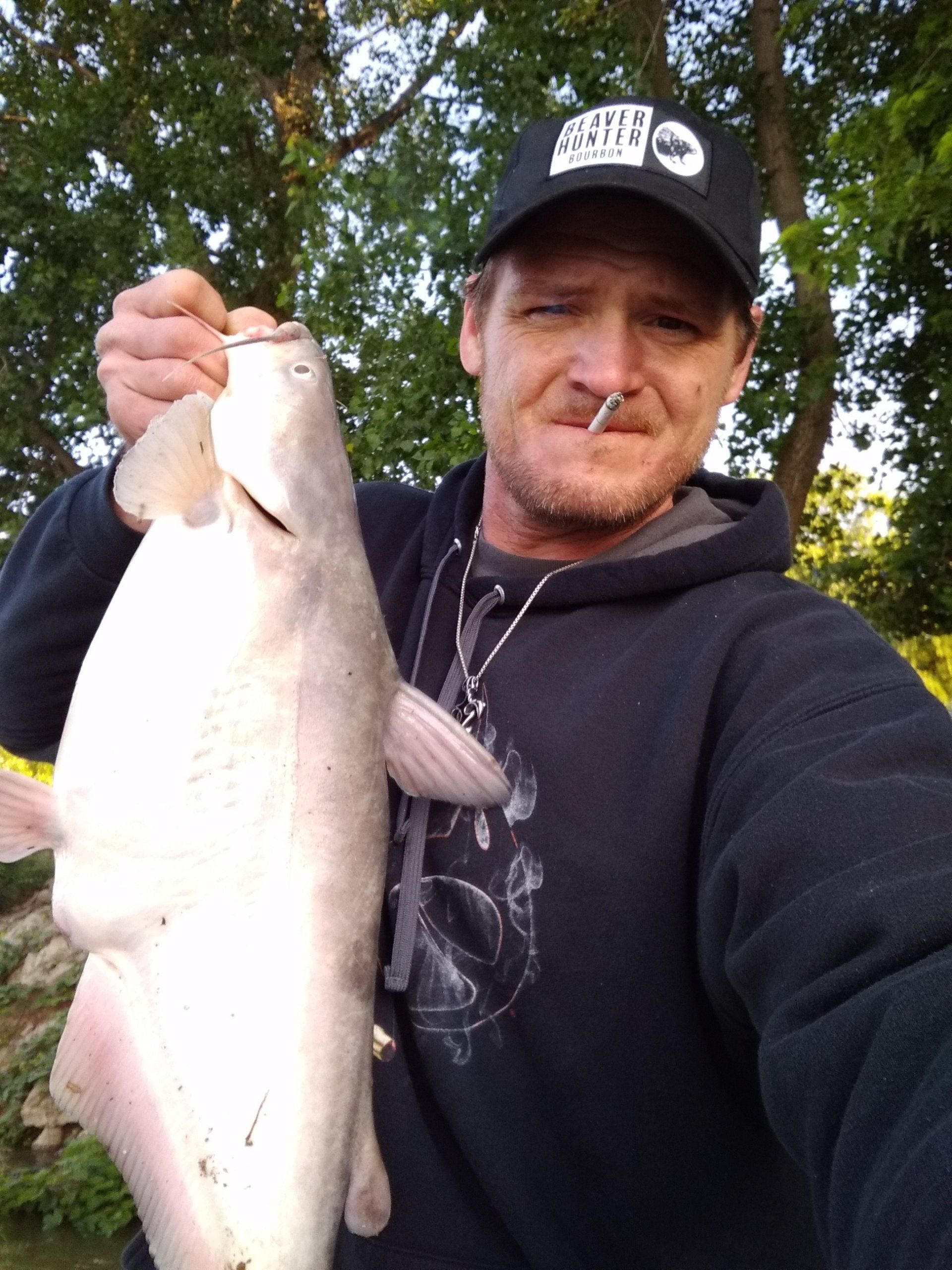 Man holding a large, light-colored catfish, wearing a black cap and hoodie outdoors.