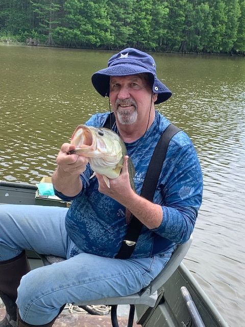 Man in a boat holding a largemouth bass he caught. Wearing a hat and blue shirt. Lake in the background.