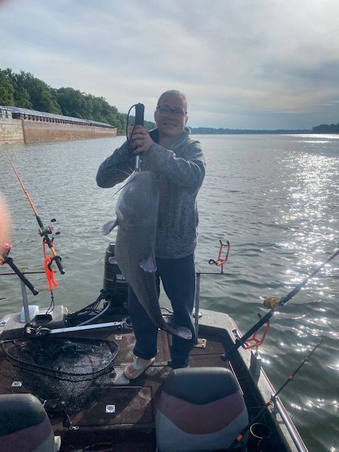 Man holding a large catfish on a boat, fishing rods visible, cloudy sky over water.