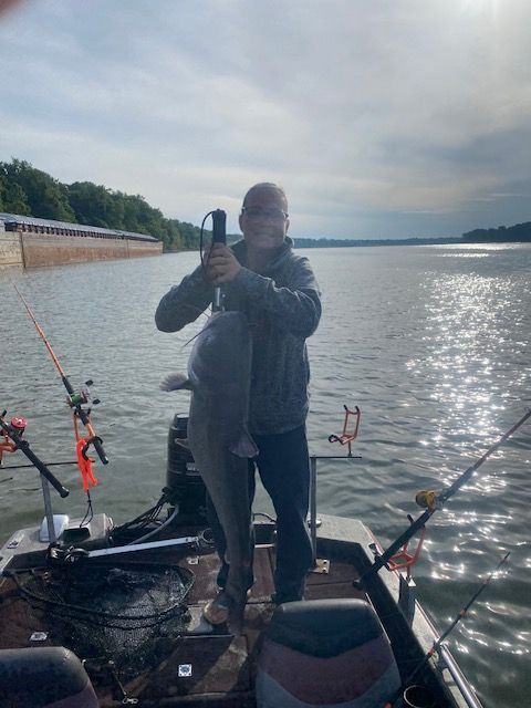 Person on a boat holding a large fish, standing in front of a wide body of water. Cloudy sky.