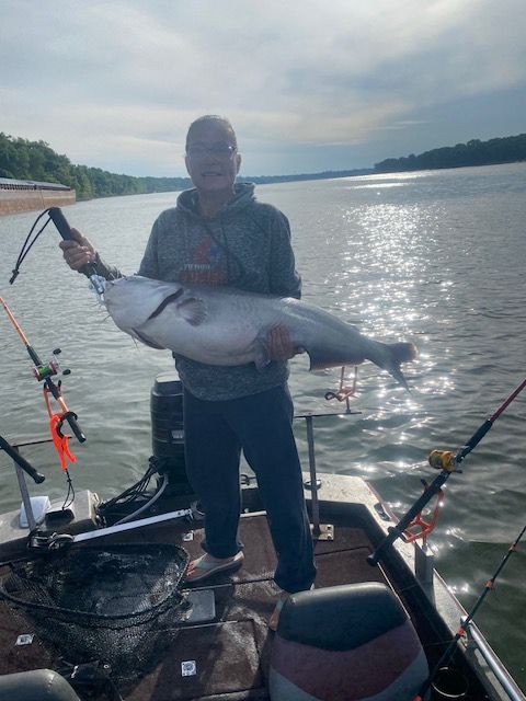 Man on a boat holding a large blue catfish, with fishing rods, and a river setting.
