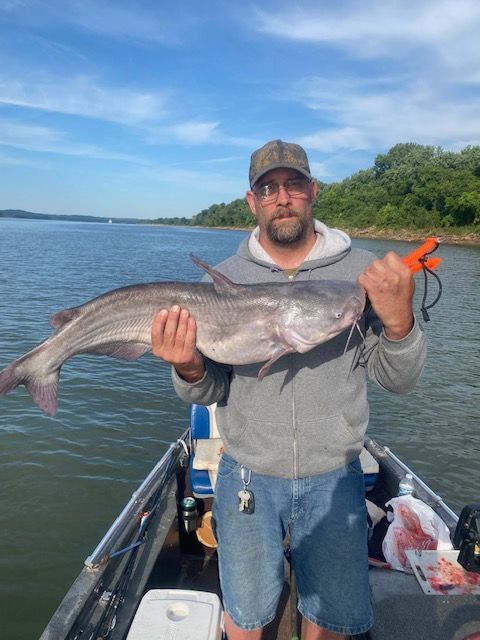 Man holding a large catfish on a boat, against a backdrop of water and sky.