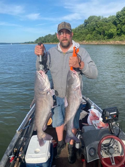 Man on a boat holding two large blue catfish he caught. River, blue sky, and trees in background.